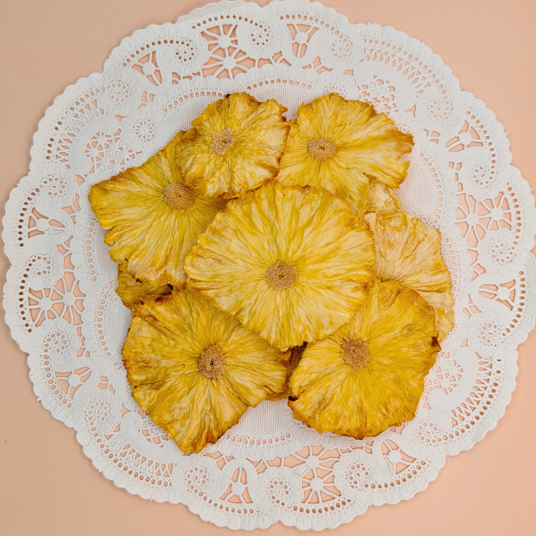 Top-down view of large golden pineapple chips artfully arranged on a white doily against a warm peach background.